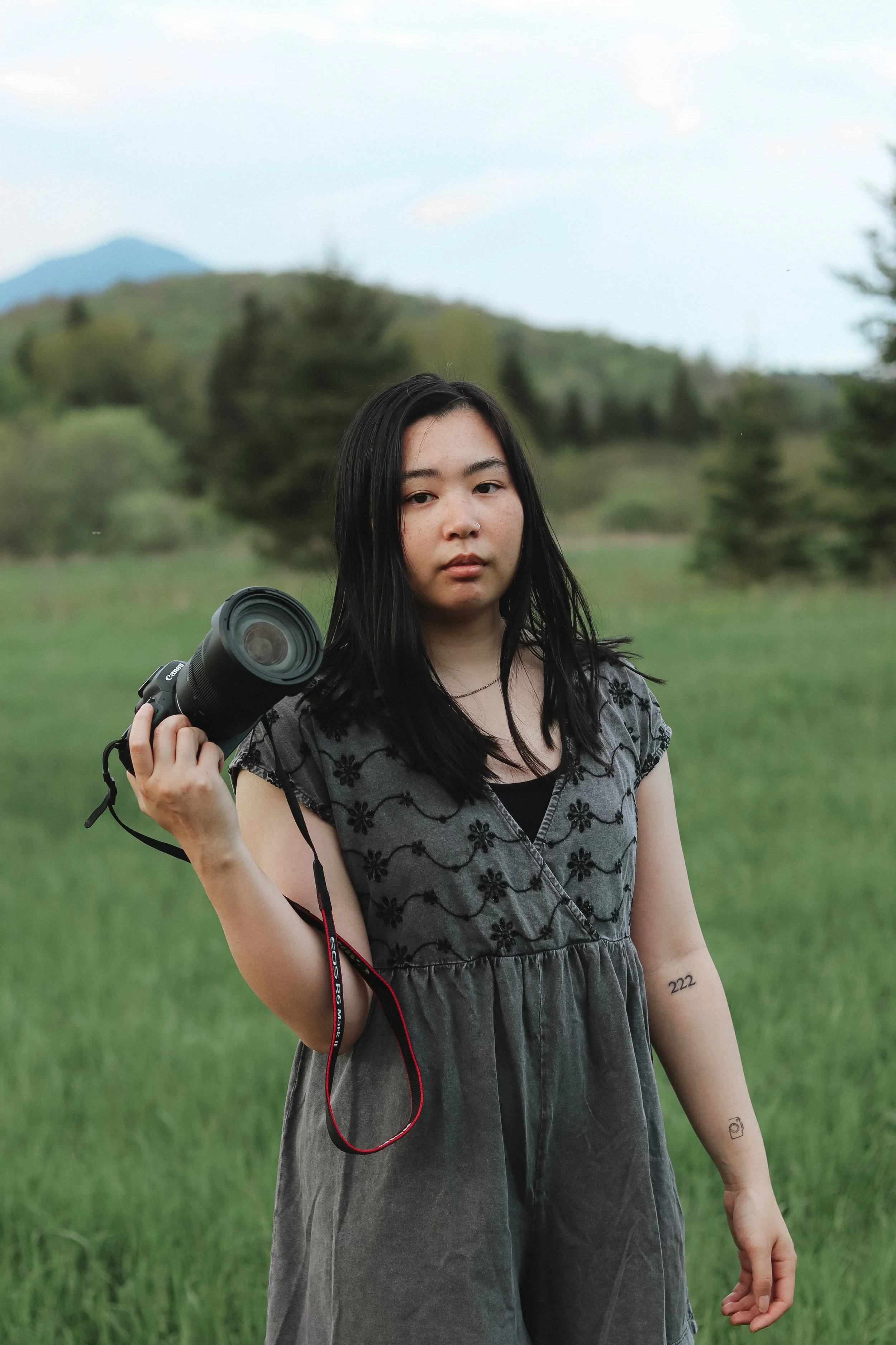 A young woman holding a camera in a grassy field with trees and mountains in the background.