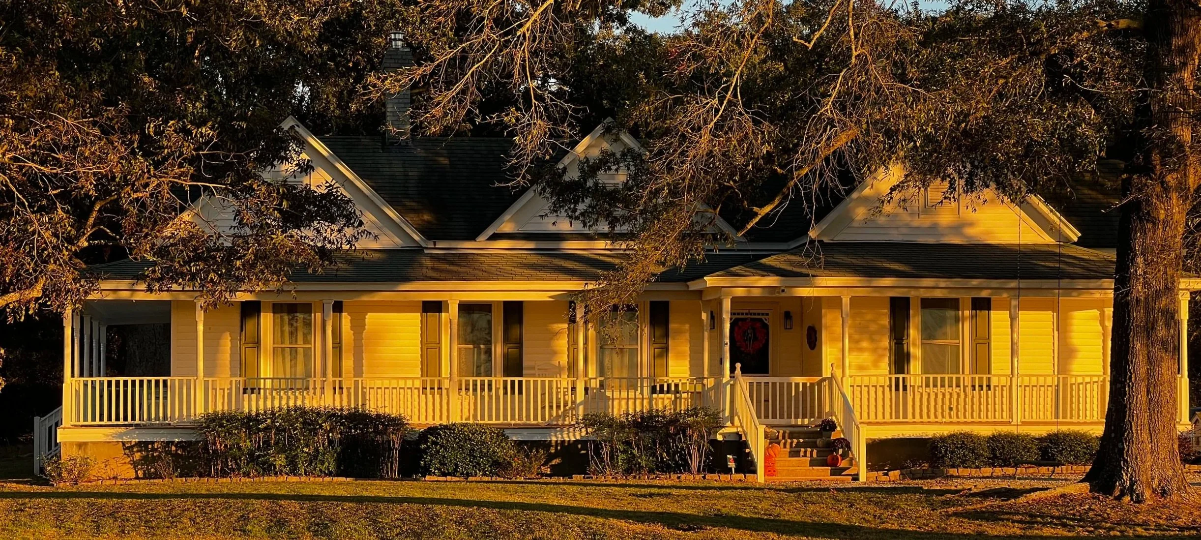 A farmhouse with a wraparound porch, dark shingle roof, front steps, and mature trees in warm evening light