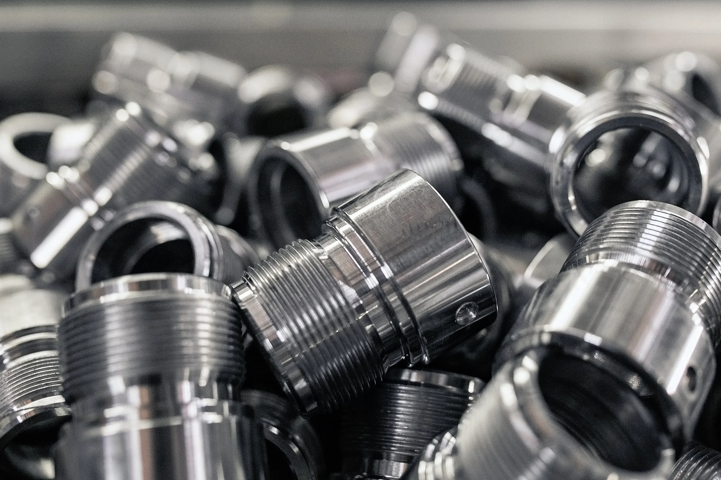 Numerous shiny, metallic, threaded pipe fittings or spacers piled together.