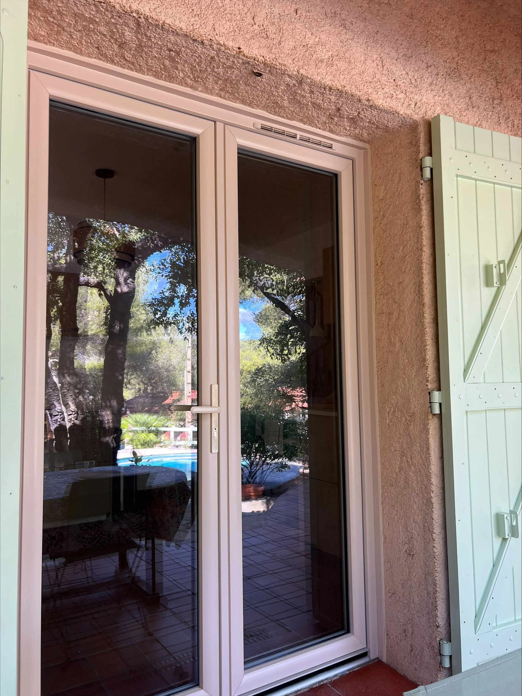 Vue d'une porte-fenêtre en verre avec volet roulant blanc, reflet de la terrasse extérieure et du jardin avec des arbres et des plantes.