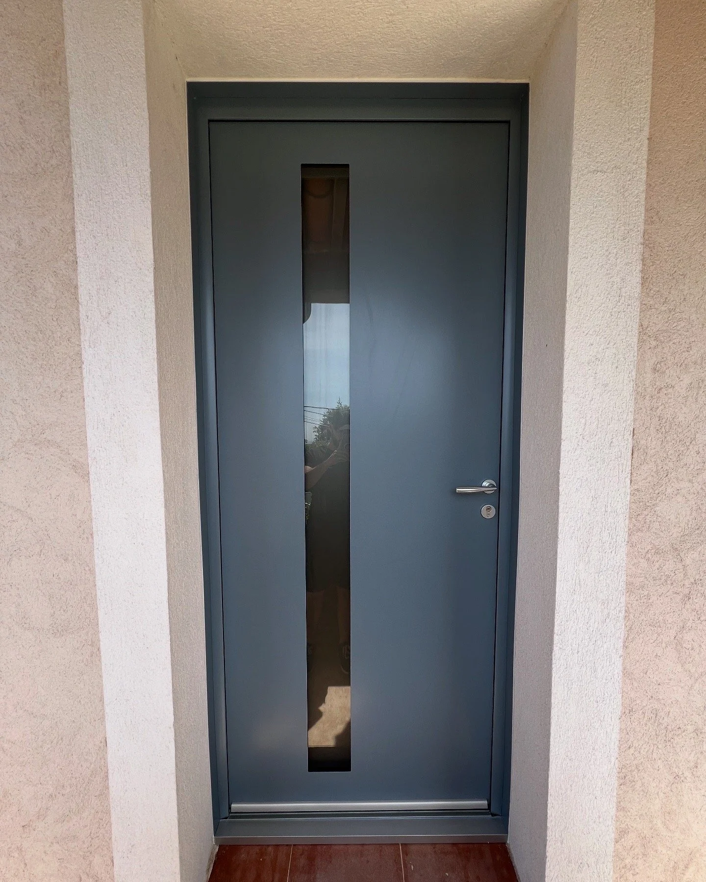 Porte d'entrée en métal de couleur gris foncé avec une vitre verticale au centre, située dans une maison.