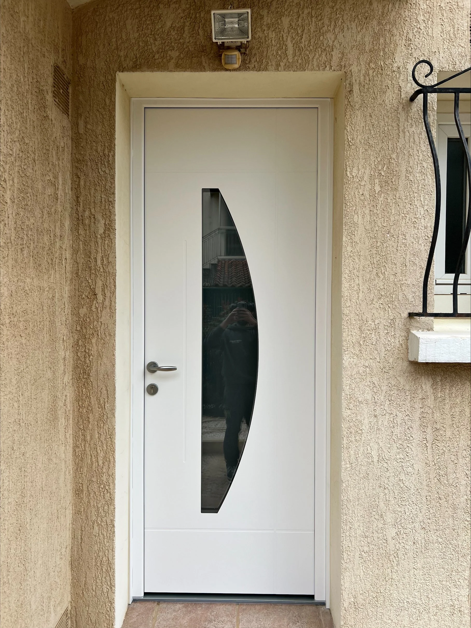 Porte d'entrée blanche avec une fenêtre en forme de goutte d'eau dans le centre, située sur un mur beige.