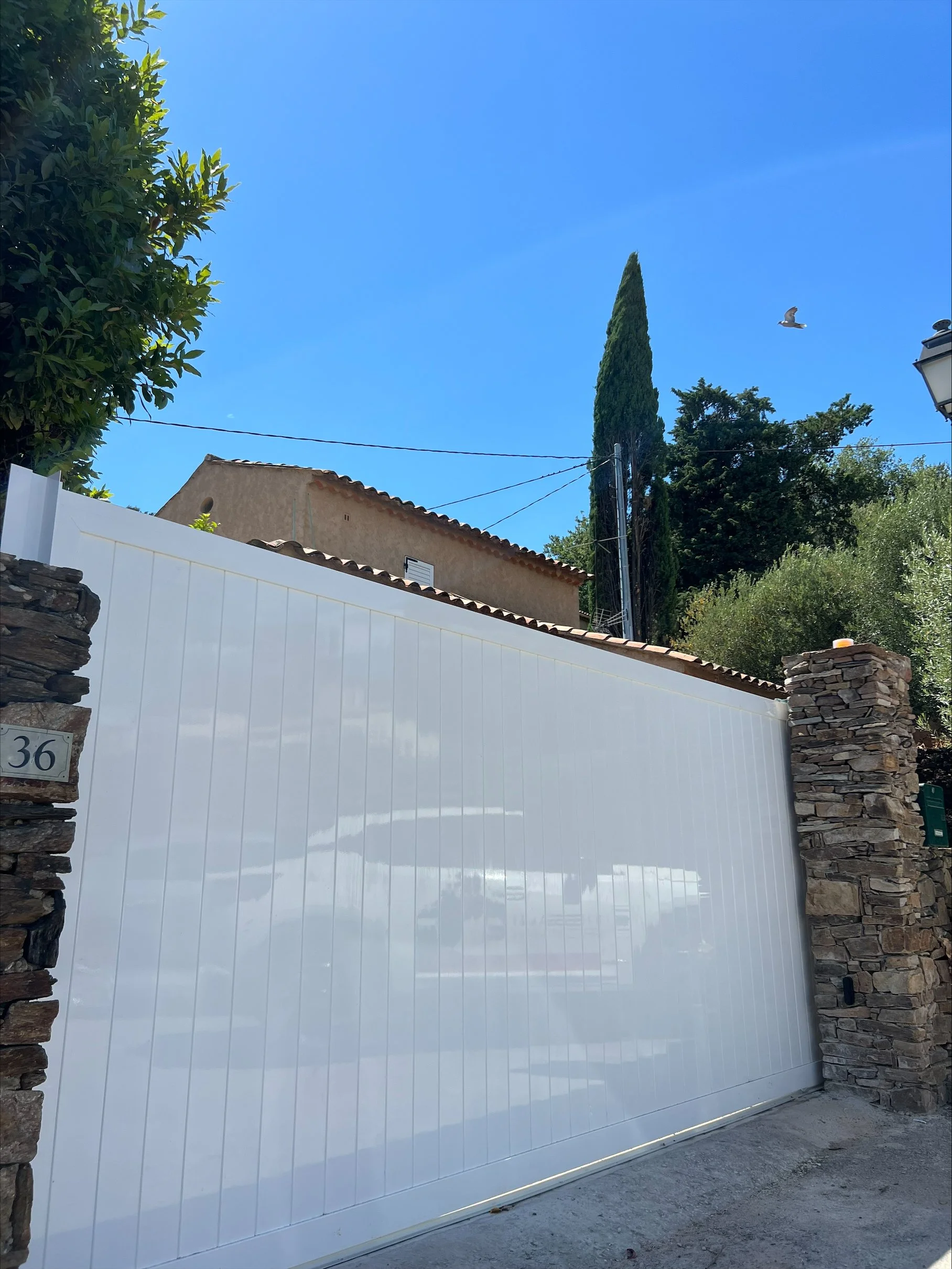 Vue d'une grande porte blanche en métal avec deux piliers en pierre, un mur en pierre à gauche, une maison avec un toit en tuiles, et un ciel bleu avec un oiseau en vol.
