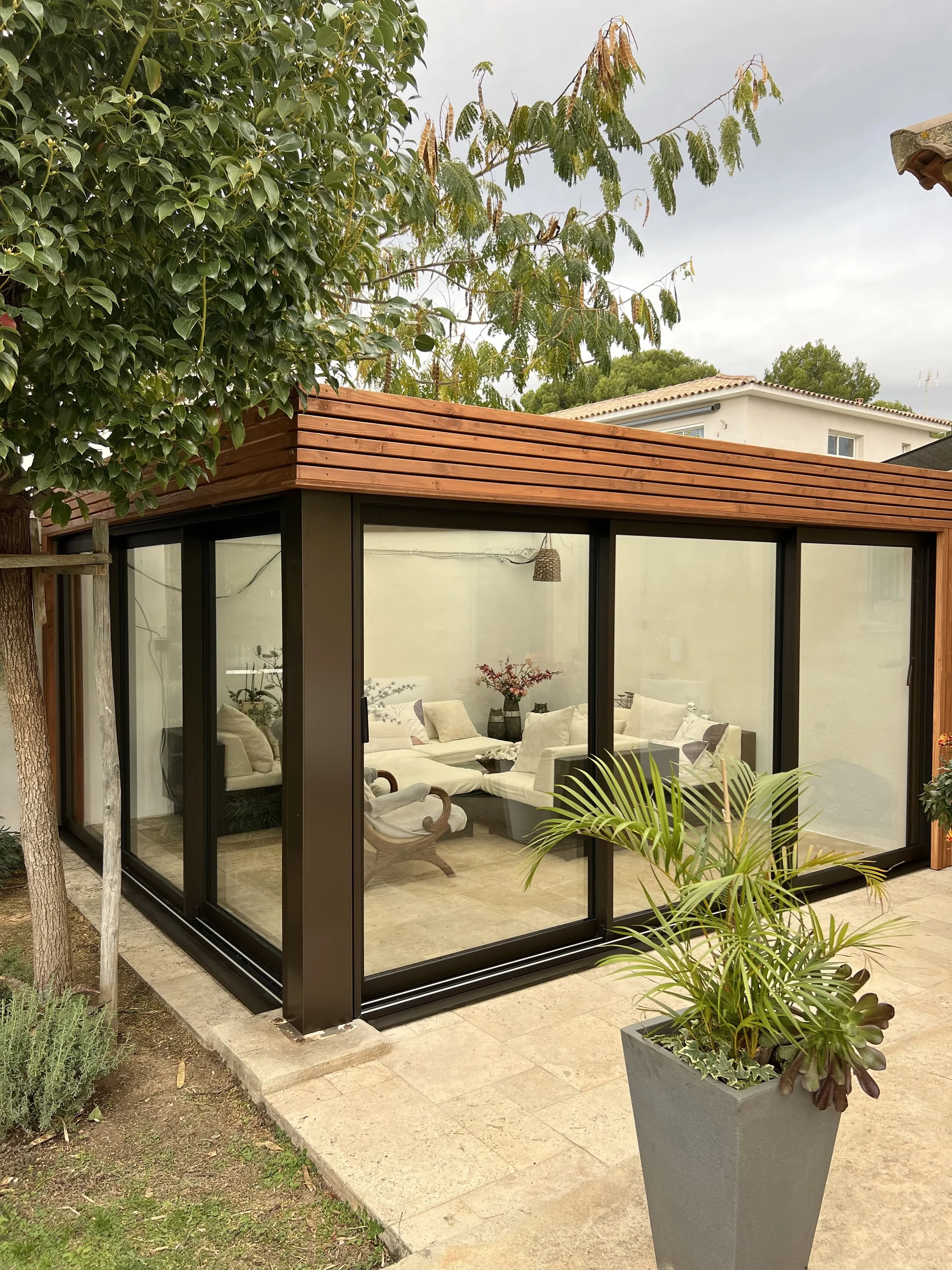 Vue d'une véranda en verre avec cadre noir, conçue pour être une pièce supplémentaire dans une maison, entourée de plantes et d'arbres, avec des meubles à l'intérieur.