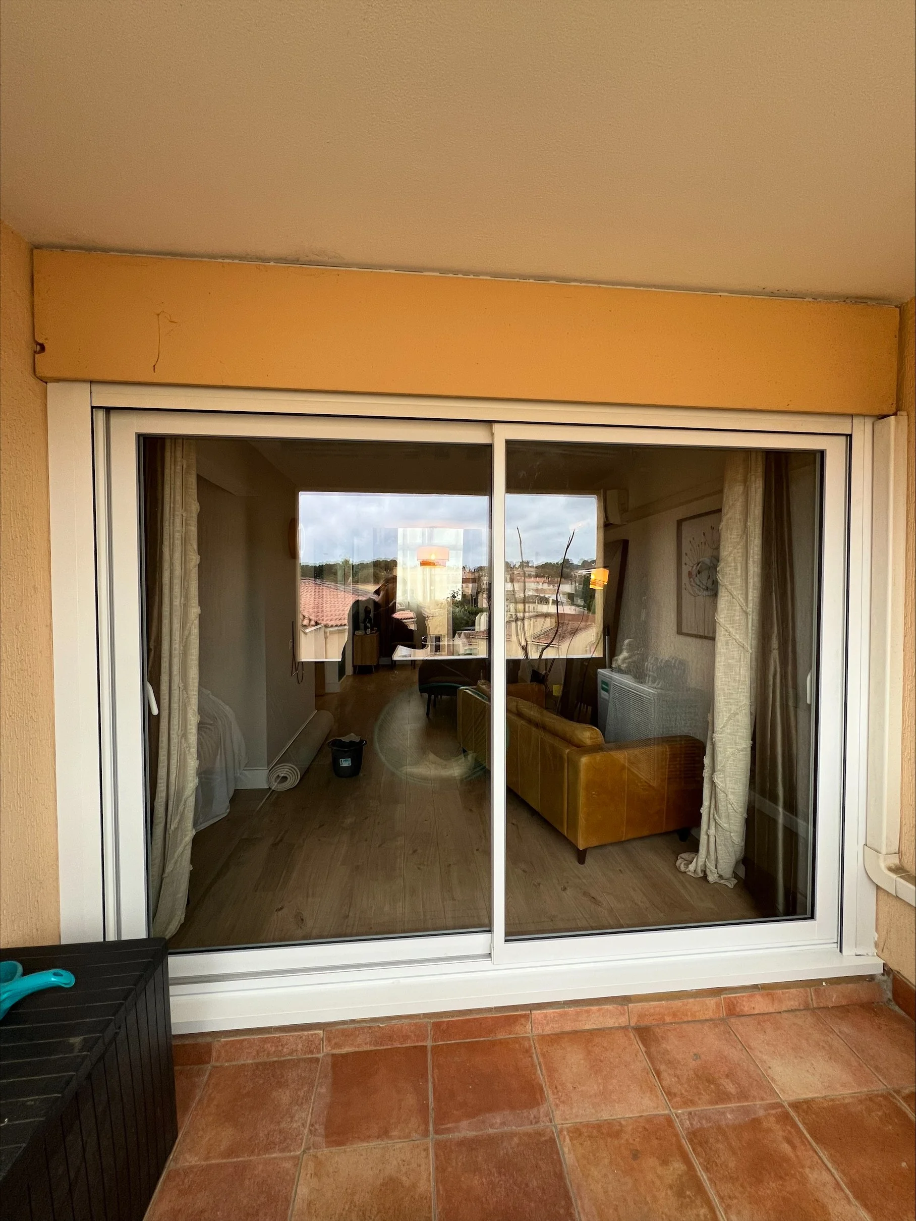 Balcon avec porte-fenêtre en verre donnant sur un salon avec canapé en cuir brun, rideaux, tableau, et vue sur des bâtiments et des arbres à l'extérieur.