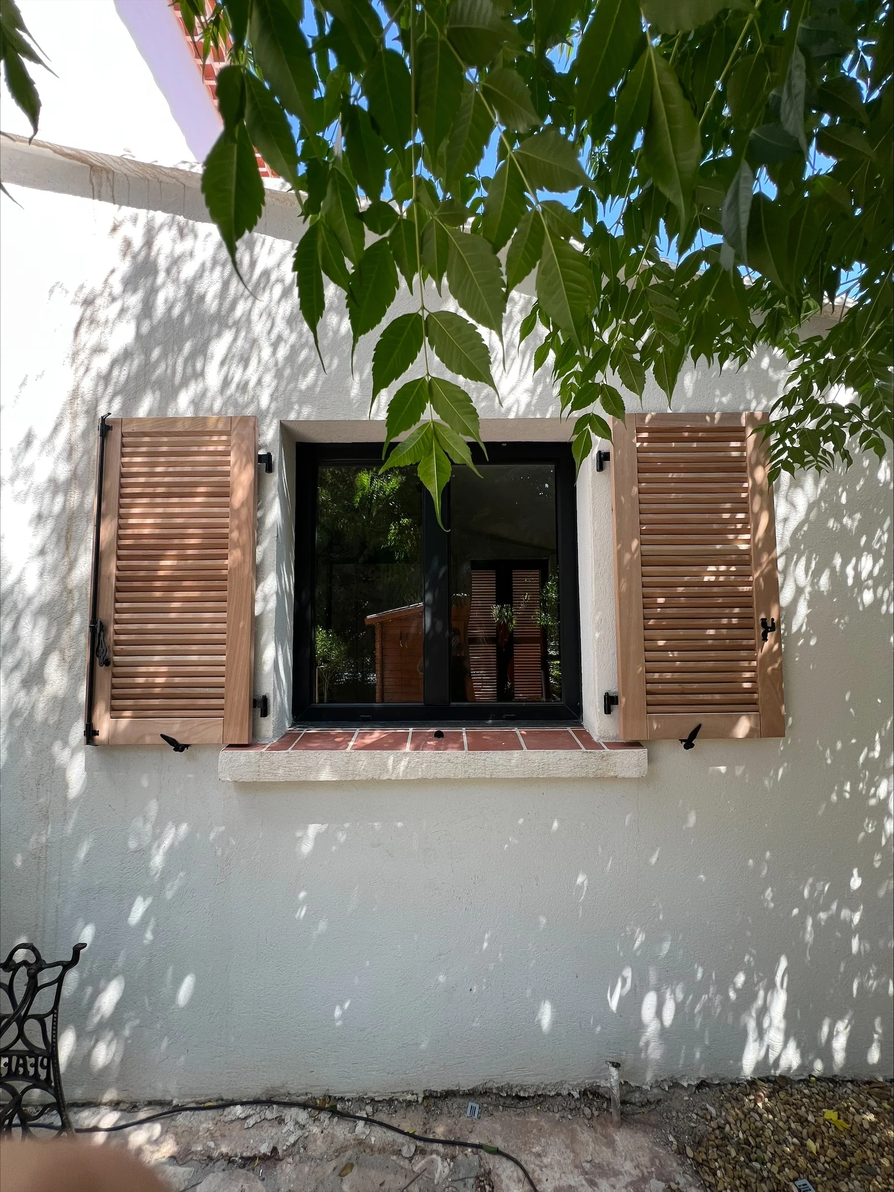 Fenêtre avec volets en bois sur un mur blanc, avec des feuilles d'arbre ombrageant la façade.