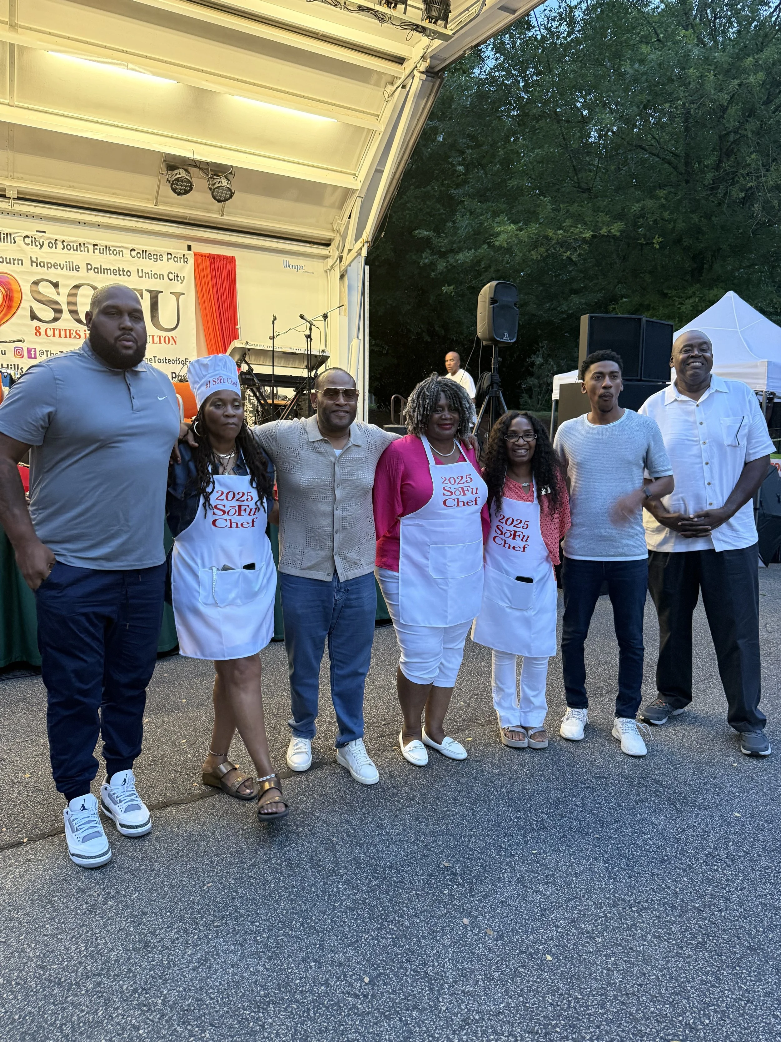 A group of seven people standing together on a stage at an outdoor event, some wearing white aprons with red text and the year 2025, with a backdrop that reads "SOFU" and lists cities, under a tent and trees.