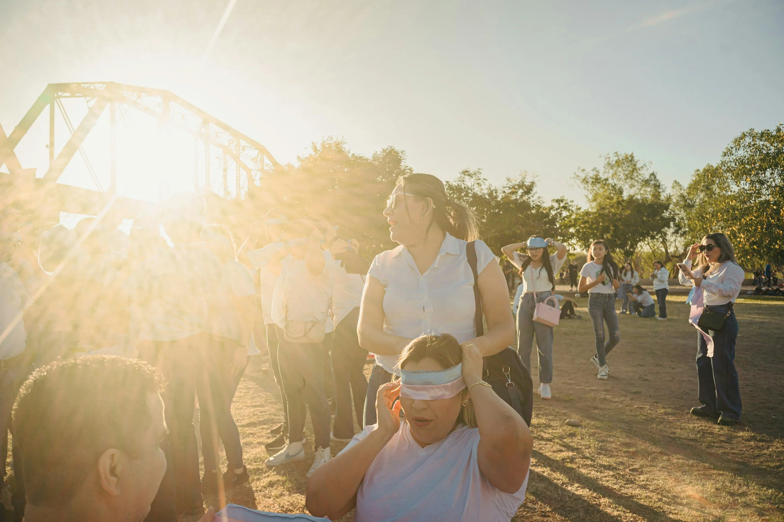 Group of people outdoors in the late afternoon or early evening, with women wearing blindfolds, participating in an activity, under the bright sun with trees and a bridge in the background.