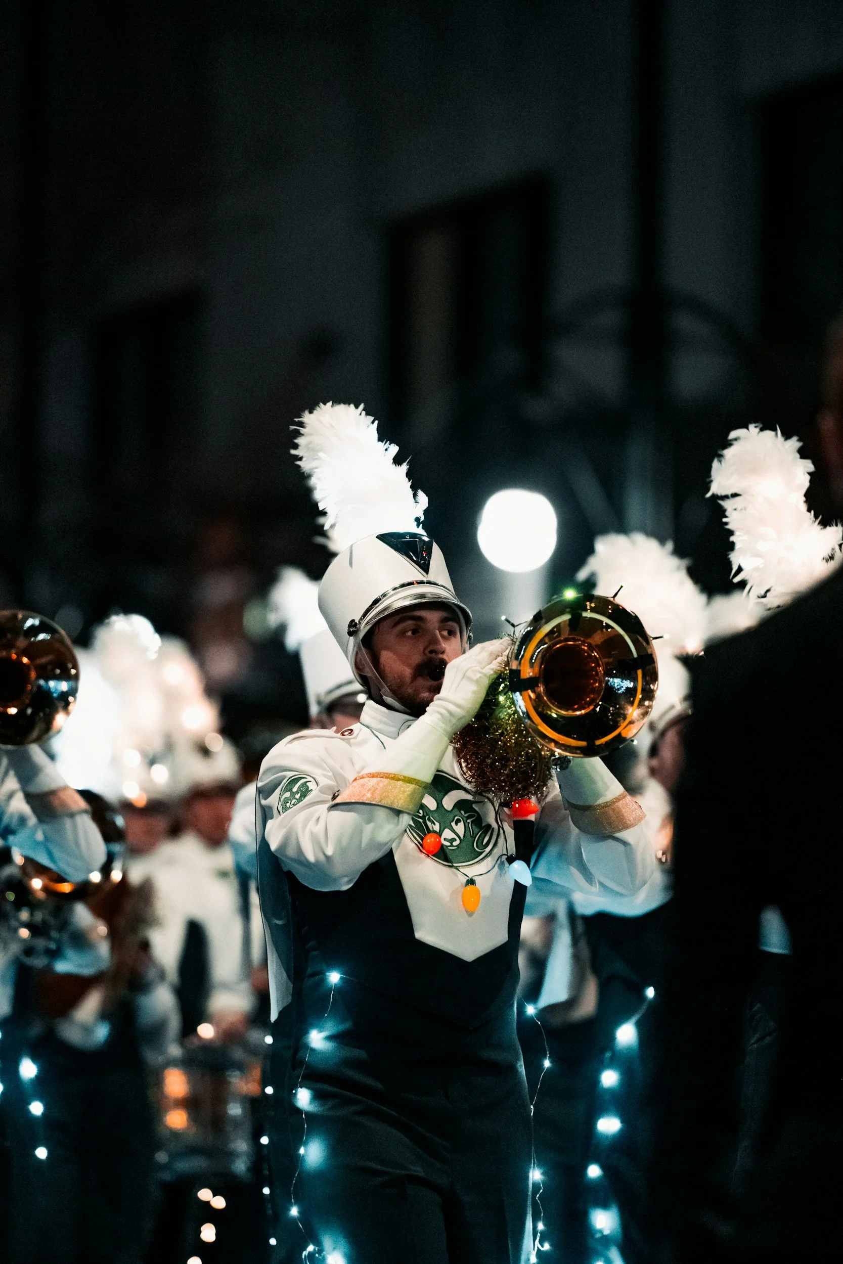 A marching band member dressed in a uniform with a white feathered hat plays a brass instrument during a night parade decorated with string lights.