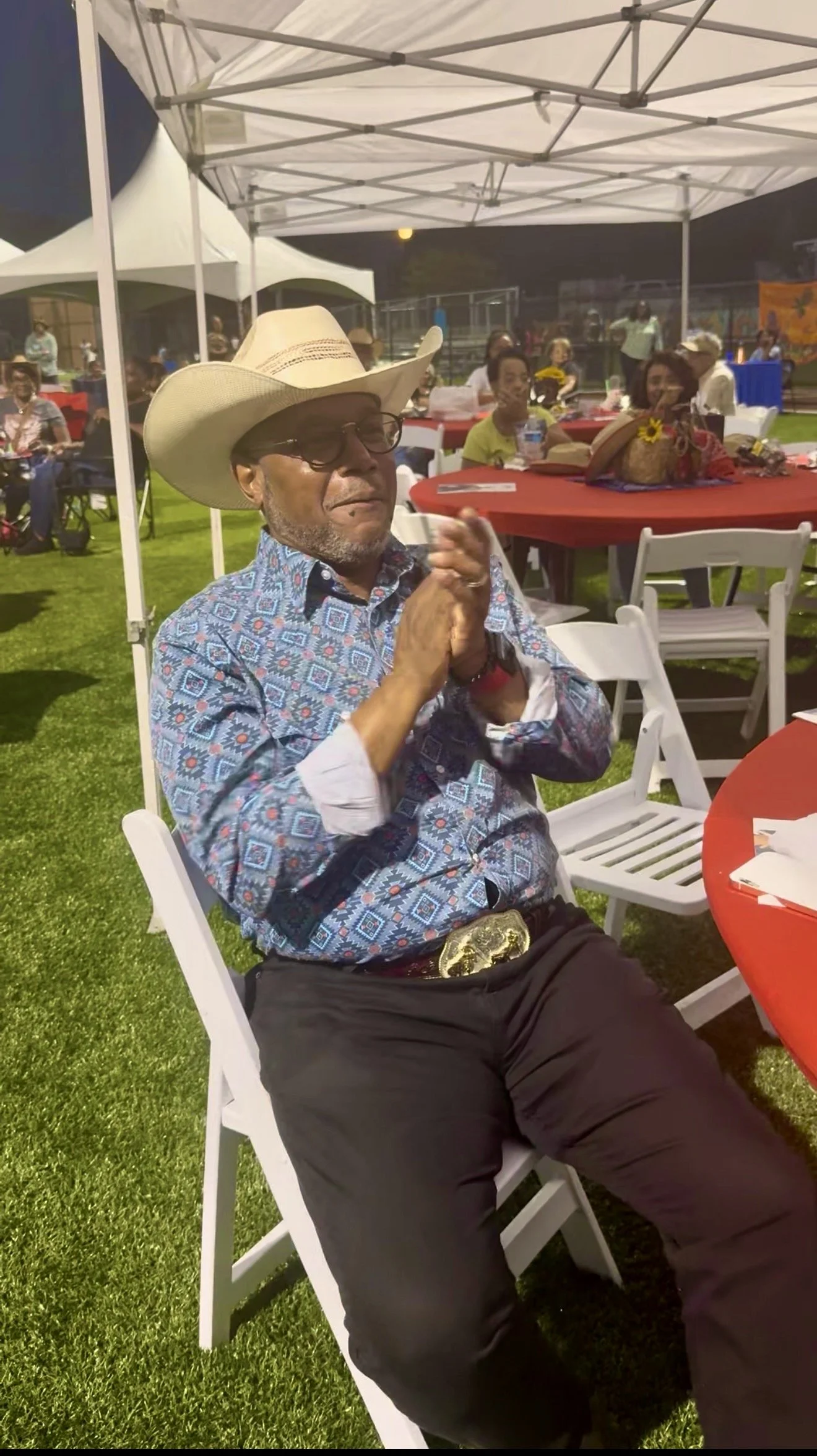 A man wearing a cowboy hat, glasses, and a patterned blue shirt sitting at an outdoor event under a white canopy, with other people sitting at tables with red tablecloths in the background.