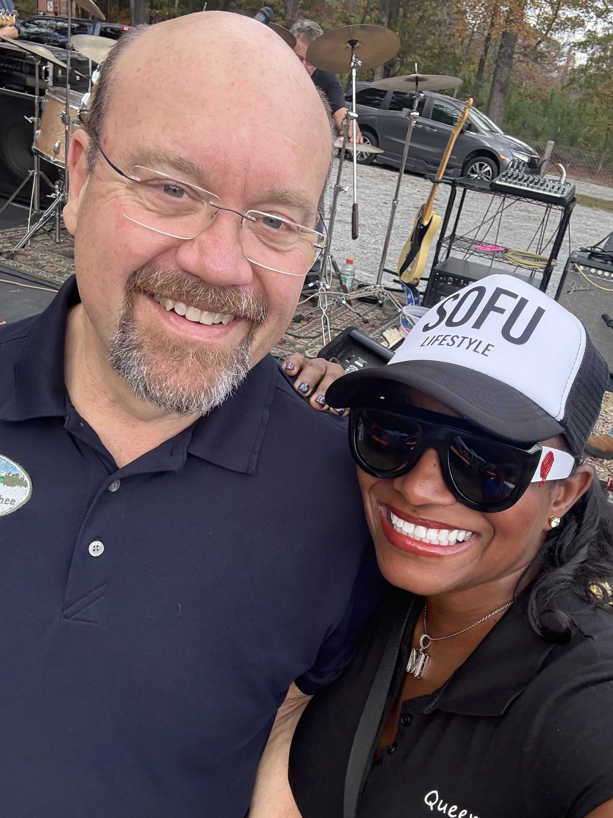 Two smiling people taking a selfie outdoors at a music event with a stage and musical instruments in the background.