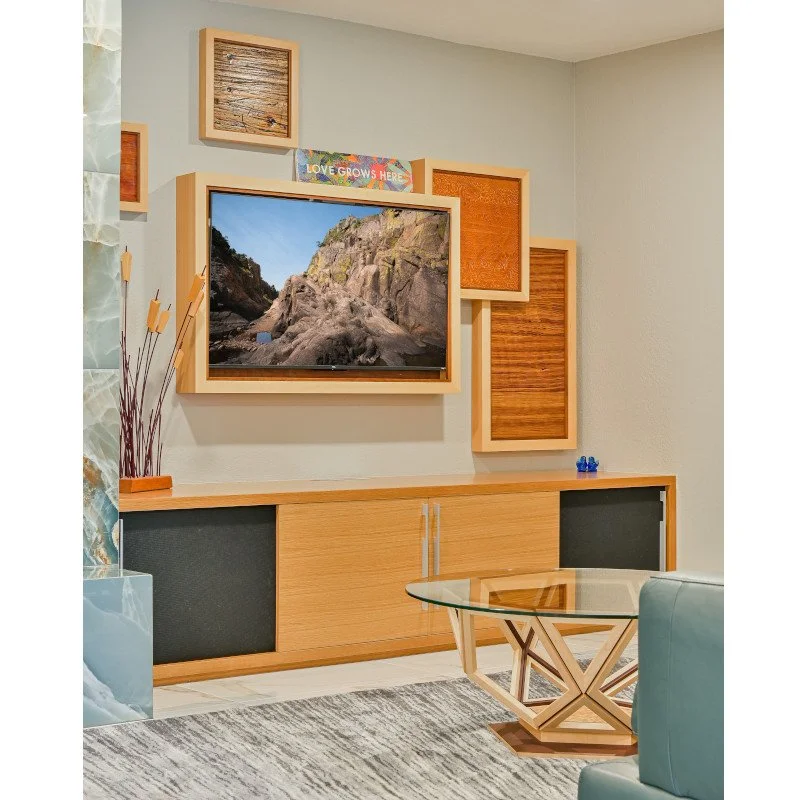 A modern living room with a wooden entertainment center and a large flat-screen TV displaying a rocky outdoor landscape. Decorative wooden frames and a colorful banner saying "Love Grows Here" adorn the wall above the TV. A glass coffee table with a 