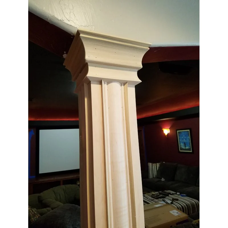 Close-up of a wooden decorative pillar in a living room setting, with a beige and brown color scheme, and a television, sofa, and framed picture in the background.