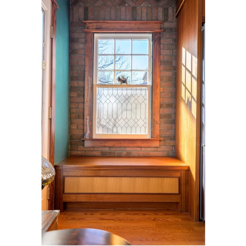 A corner of a room with a large window featuring decorative glass, surrounded by wood trim, with brick wall behind it and wooden paneling on the right side. Sunlight casts a shadow of the window on the paneling.