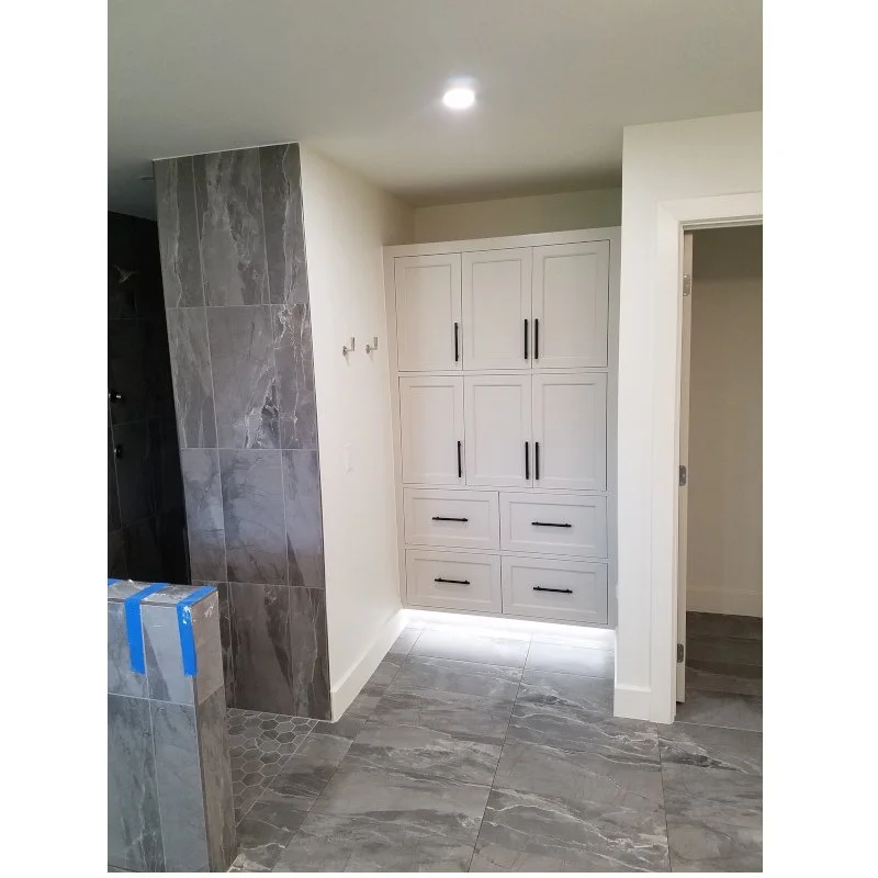 A corner of a modern bathroom with gray marble floor tiles, a white built-in cabinet with black handles, and a gray tiled wall near a shower area.
