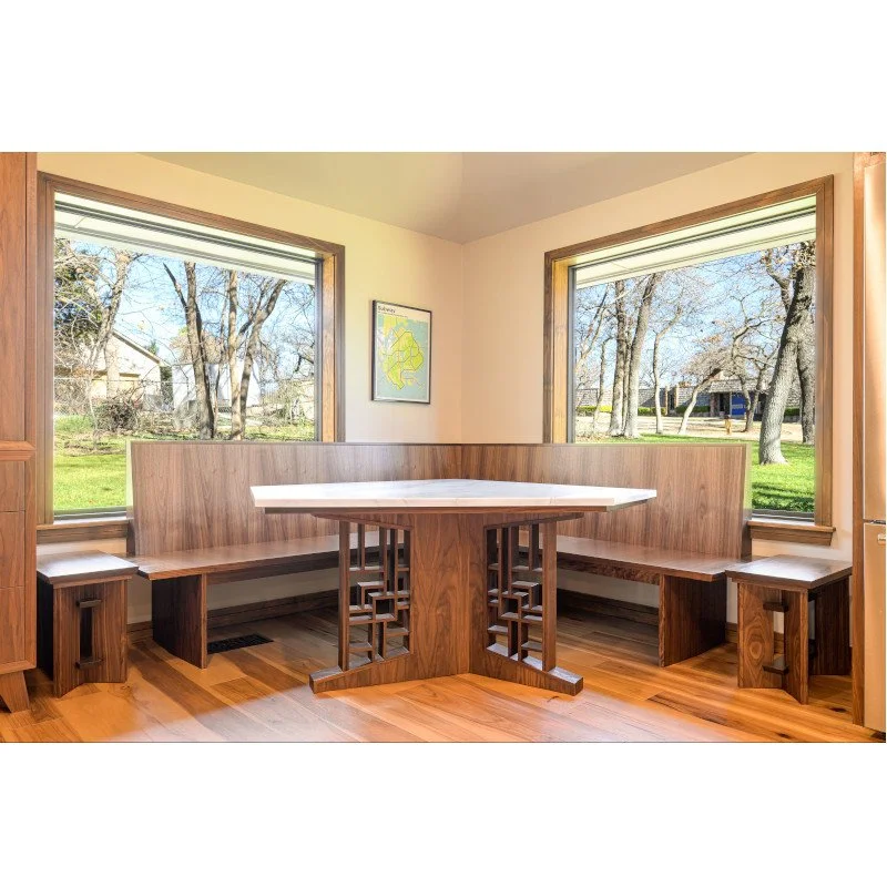 A corner nook with a wooden built-in booth seating area, asymmetrical dining table, and two small stools, surrounded by large windows showing an outdoor yard with trees and a house.