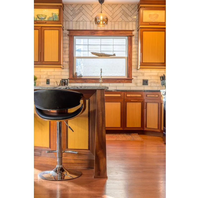 A cozy kitchen featuring wooden cabinets, a window with a fish-shaped decor, a black swivel stool, a granite countertop, and a brass pendant light.
