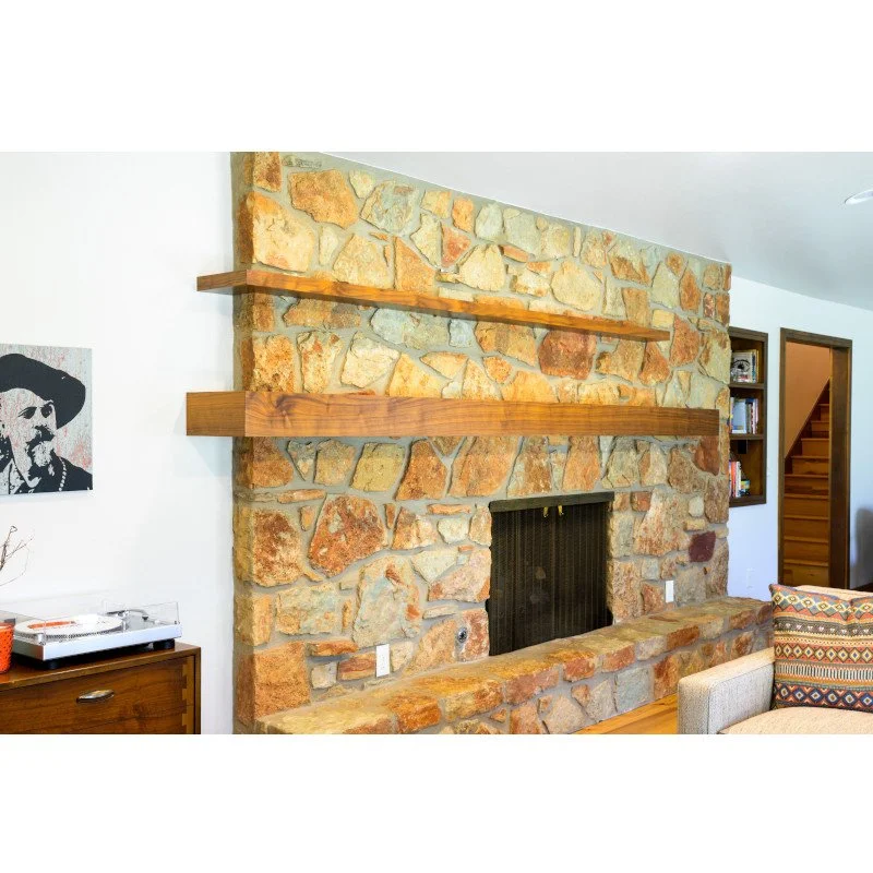 Living room with a large stone fireplace, two wooden shelves above the fireplace, a colorful patterned pillow on a beige armchair, and a black and white portrait on the wall.