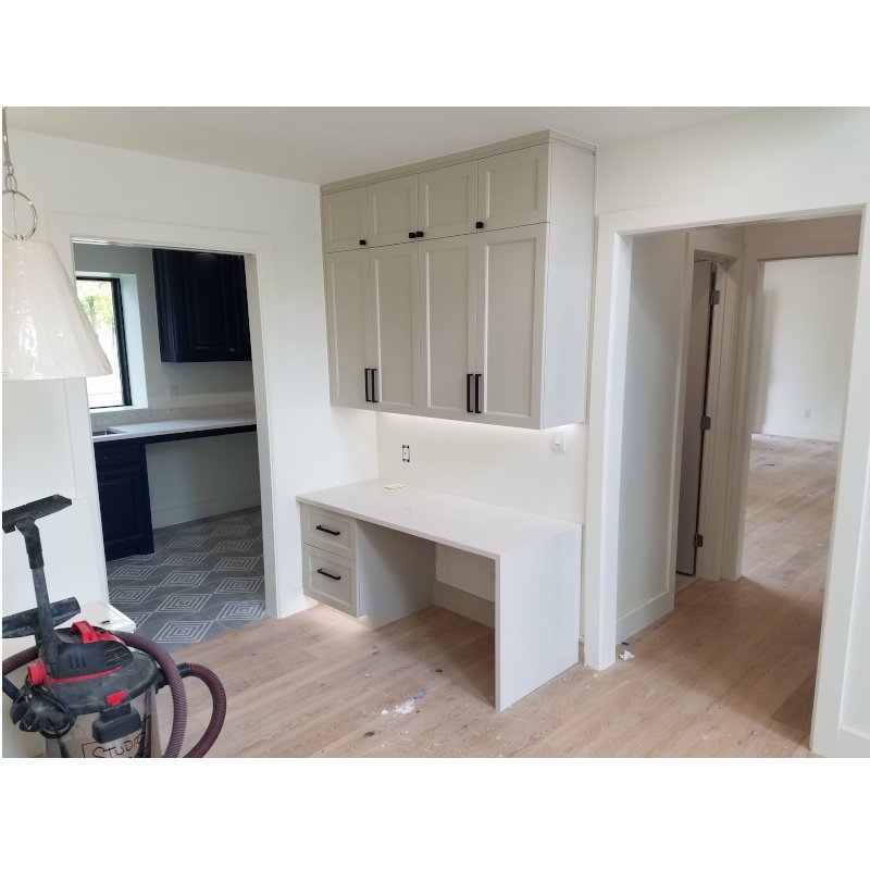 Room with white cabinets and desk, open doorways to kitchen with dark cabinets and an adjacent room, vacuum cleaner on hardwood floor.