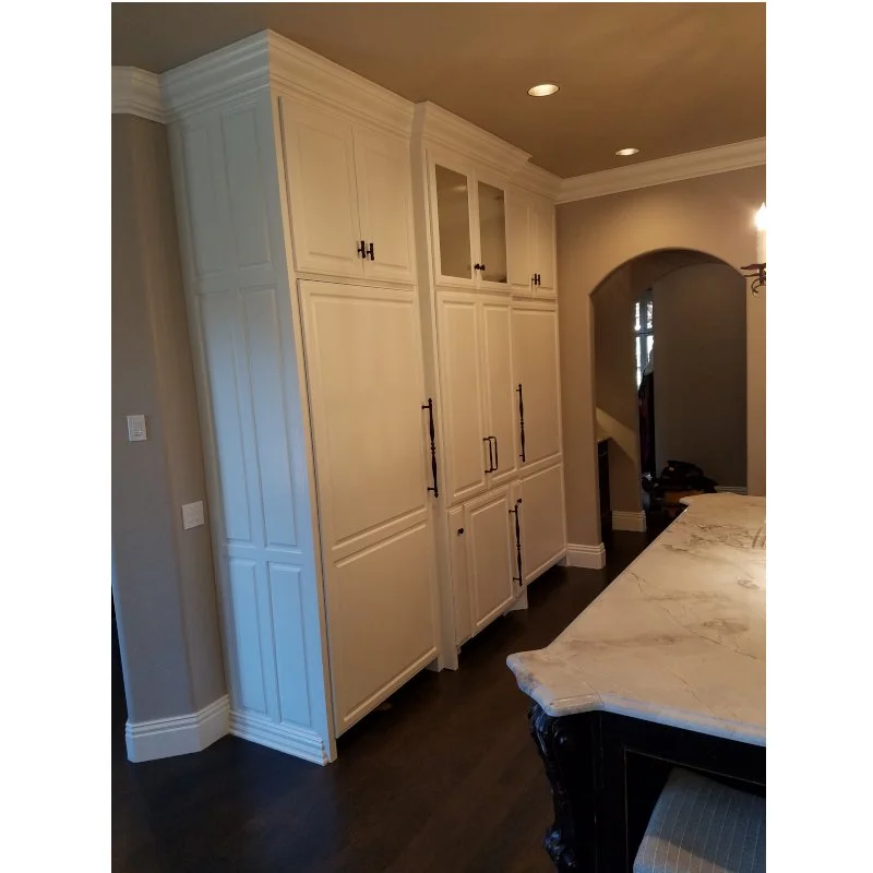 White built-in kitchen cabinets with black handles, next to a kitchen countertop with a marble surface, in a home interior with dark hardwood floors and an arched doorway.