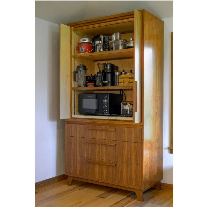 A wooden kitchen cabinet with open shelves, containing a red slow cooker, blender, coffee grinder, and various small appliances, with a microwave oven on the built-in countertop.