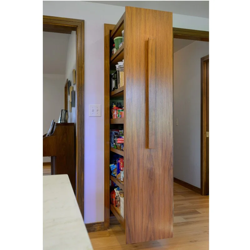 A tall wooden pantry pull out unit with various snacks, viewed from a dining area, with doors and wall outlets visible in a home interior.