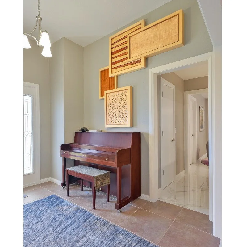 An upright wooden piano with a matching bench in a corner of a room, with a modern abstract wall art piece above it, near a doorway and a window with decorative glass, in a well-lit space.
