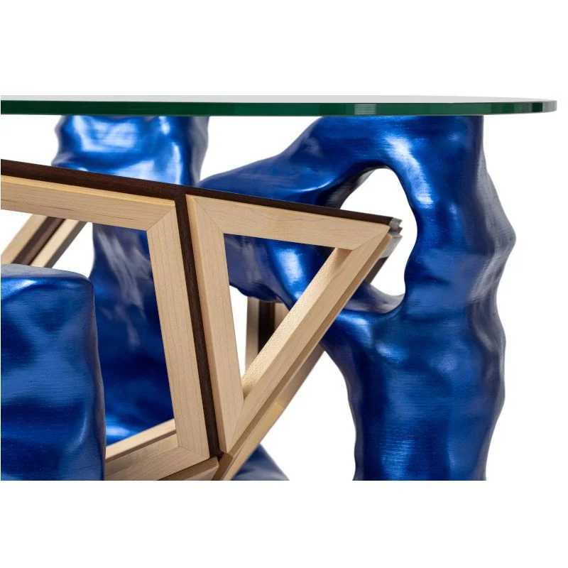 Close-up of a uniquely designed table with a glass top, blue textured sculptural members, and a wooden frame with geometric shapes.