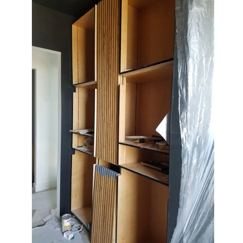 A tall wooden shelving unit with multiple open compartments, some filled with papers and a few tools, against a dark wall, with a plastic sheet partially covering the right side.
