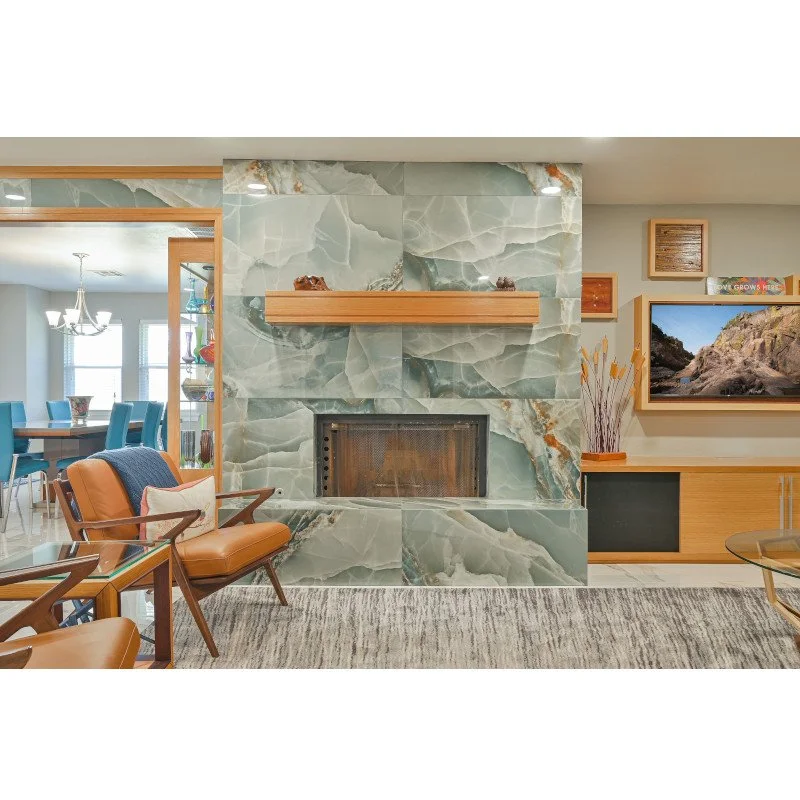 A mid-century modern living room with a large green marble fireplace, tan leather armchairs, a wooden cabinet, and a wall-mounted flat-screen TV.