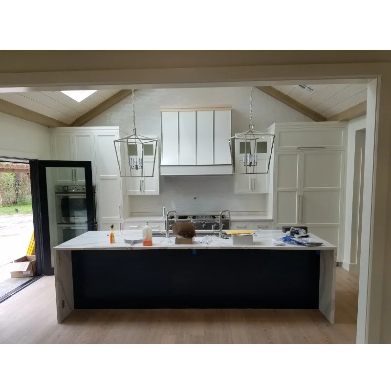 Modern kitchen with white cabinetry, a central island with a dark blue front, stainless steel appliances, and two geometric pendant lights. The room is bright with natural light from sliding glass doors.