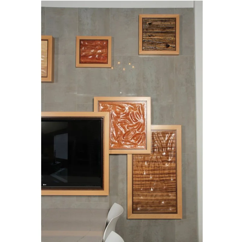 Multiple framed wooden art pieces hanging on a gray tiled wall, with a large black television screen partially visible in the lower left corner and a white chair at the bottom.