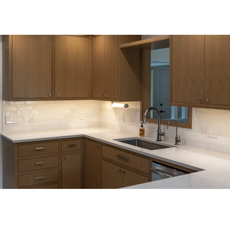A modern kitchen with wooden cabinets, white countertops, a stainless steel sink, and a window above the sink. Under-cabinet lighting and a soap dispenser are also visible.