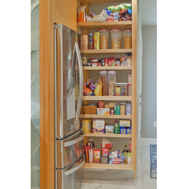 Open slide out pantry with wooden shelves filled with snacks, cereals, and jars, next to a stainless steel refrigerator in a kitchen.