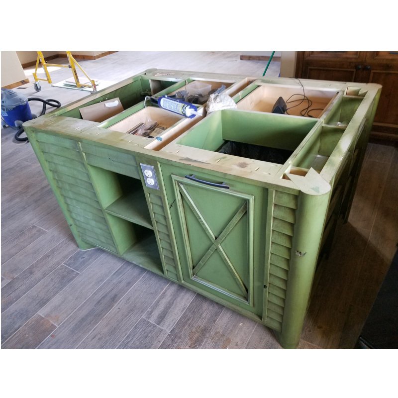 Green wooden kitchen island under construction with open compartments and a closed cabinet door, placed on a wooden floor.