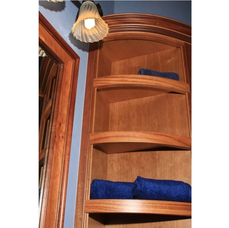 A corner wooden bookshelf with three shelves, two of which are holding dark blue towels, and a wall-mounted lamp with a glass shade above.