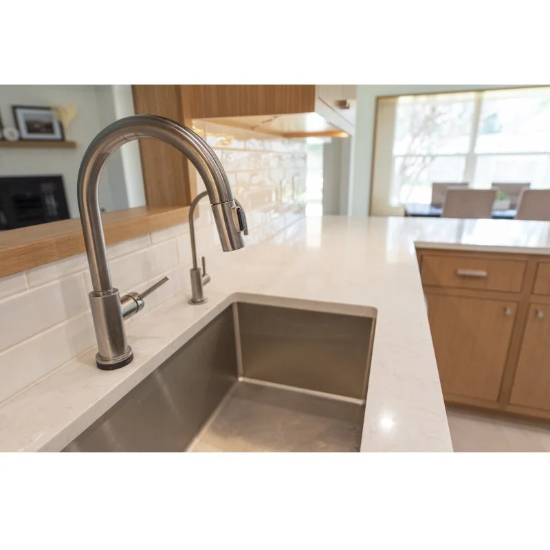 Close-up of a modern kitchen sink with a stainless steel faucet and wood accents, overlooking a breakfast area with large windows.