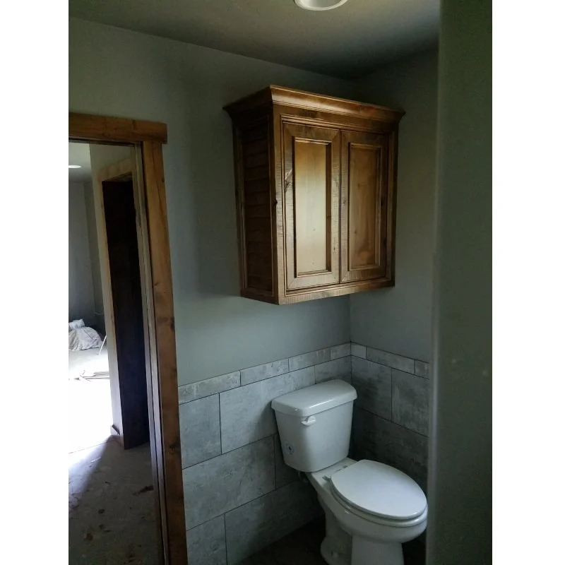 Bathroom with a white toilet and wooden cabinet above it, partial view of a room through a doorway, and tiled wall behind the toilet.