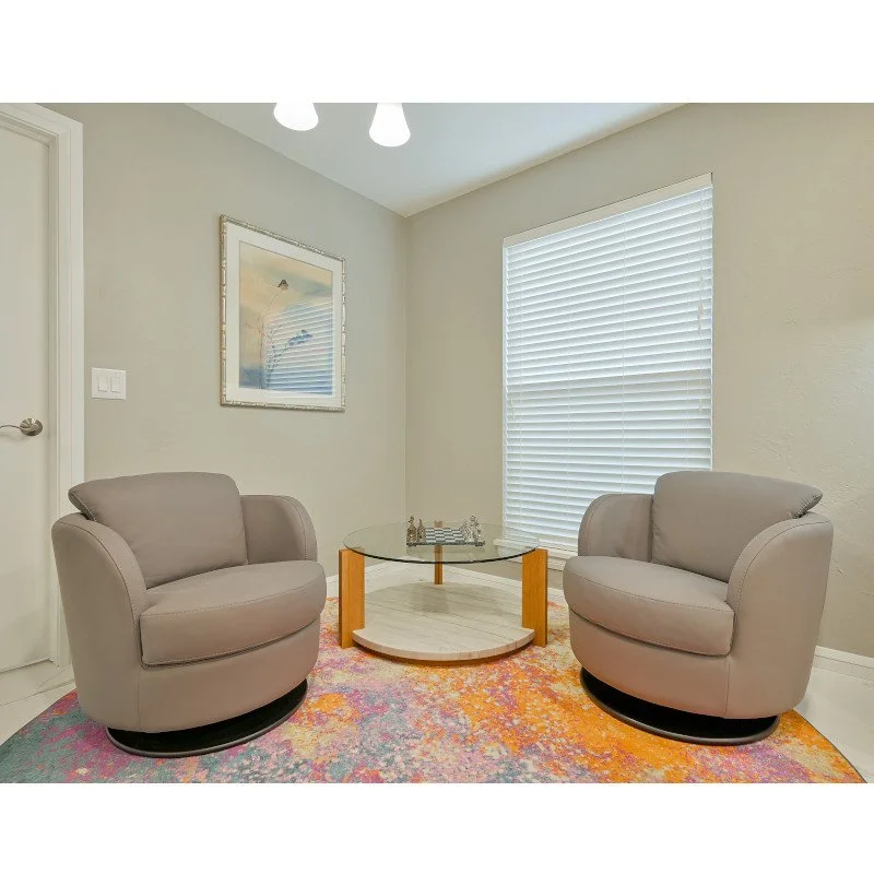 A small living room corner with two beige armchairs, a glass coffee table, a colorful rug, a window with white blinds, and a framed picture on the wall.