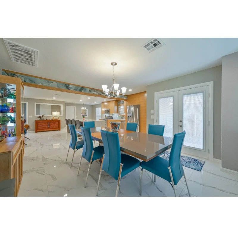 A modern dining area with a rectangular table and blue chairs, marble flooring, and a glass door leading outside. In the background, a kitchen with appliances and a small living area.