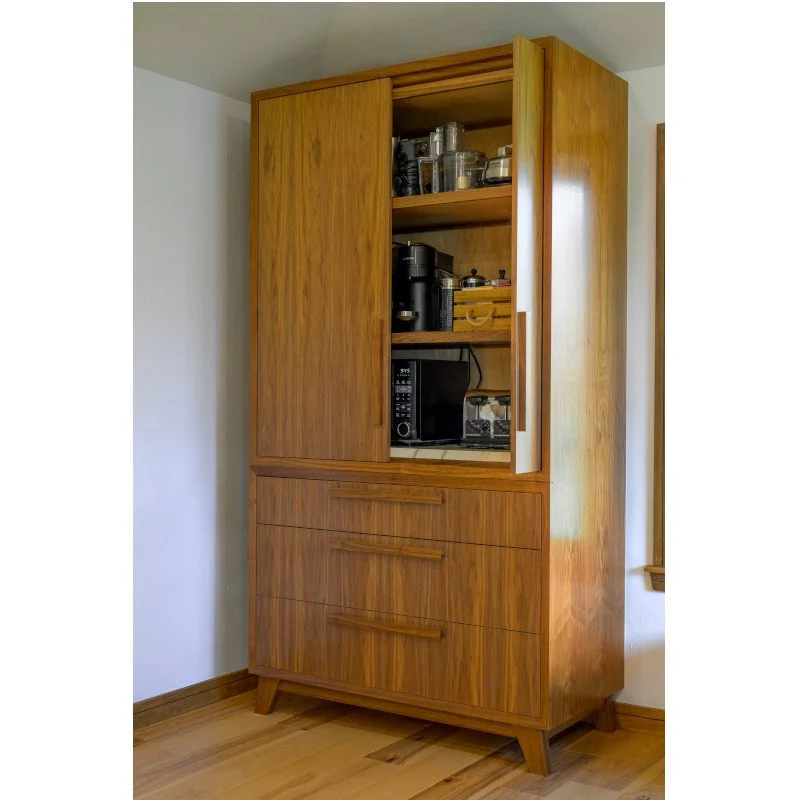 Wooden kitchen cabinet filled with kitchen appliances and glassware, with closed and open doors, standing on a hardwood floor next to a white wall.