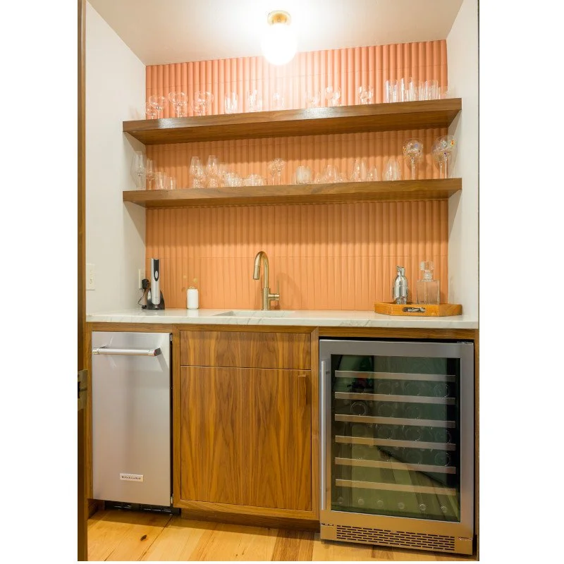 Home bar with wooden cabinetry, a marble countertop, open wooden shelves with wine glasses, a small wine fridge, and  coral-colored textured wall tile.