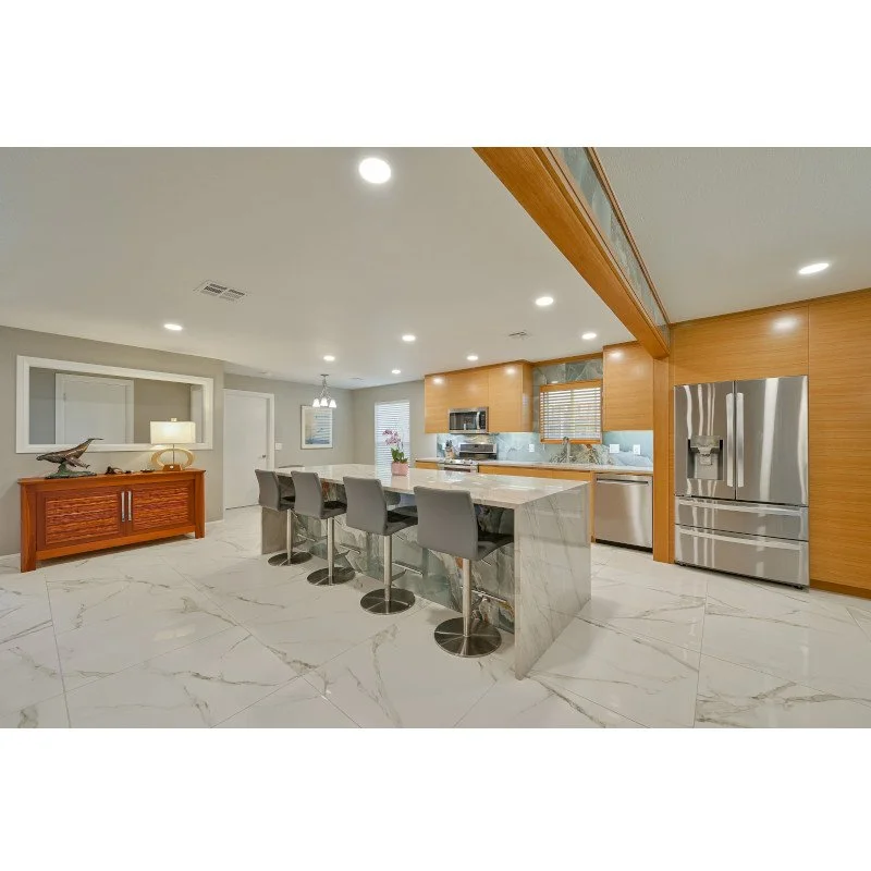 Modern open-concept kitchen with marble flooring, wooden cabinets, and stainless steel appliances, including a refrigerator; kitchen island with seating and a gray chairs; adjacent dining area with table and chandelier; decorative items and windows w