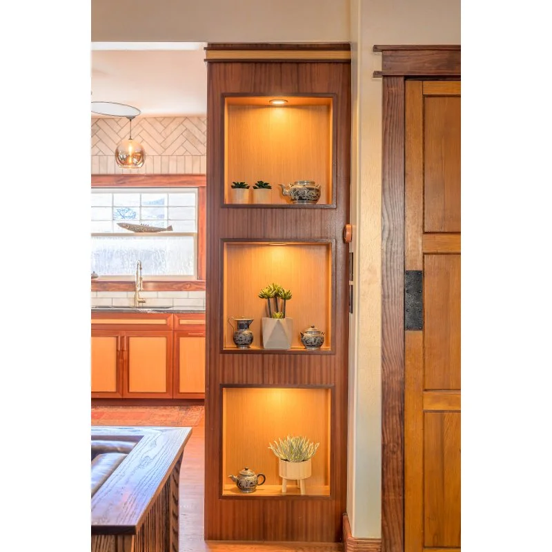 A vertical wooden display shelf with three illuminated niches holding small potted plants and decorative objects, located in a kitchen or dining area with wooden cabinets and a window in the background.