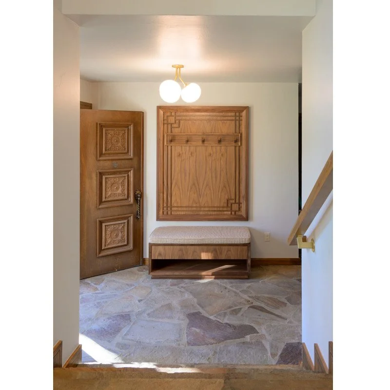 An entryway with a wooden door, a built-in wooden hall tree with hooks, a cushioned bench underneath, and a stone floor. A ceiling light fixture with two globes illuminates the area.