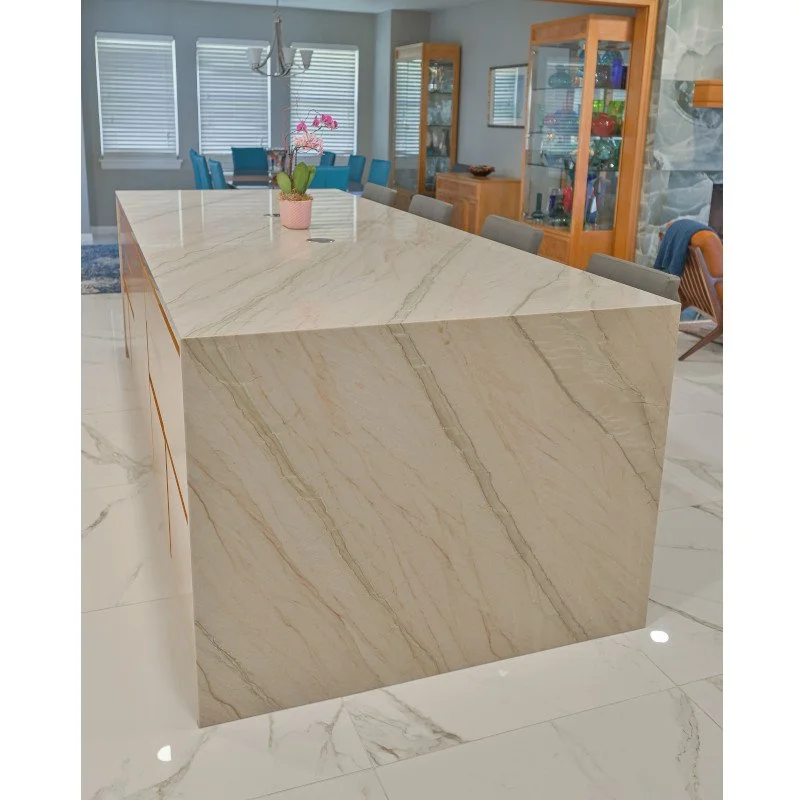 A modern kitchen island made of light-colored quartzite with natural veining, situated in a dining area with a chandelier, blue dining chairs, and wooden display cabinets in the background.