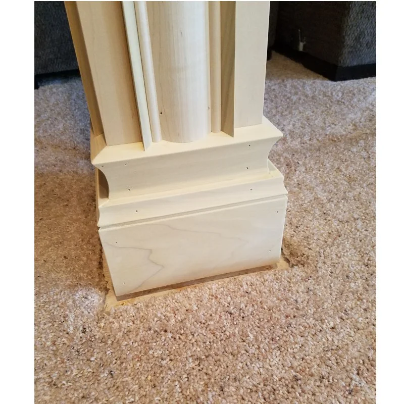 Close-up of a wooden column foot, placed on beige carpet, with part of a dark-colored sofa visible in the background.