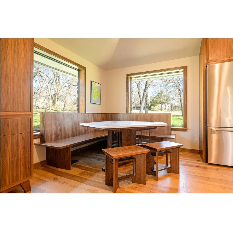 A kitchen corner with a wooden dining table and benches, large windows showing trees outside, a stainless steel refrigerator, and wooden cabinetry.