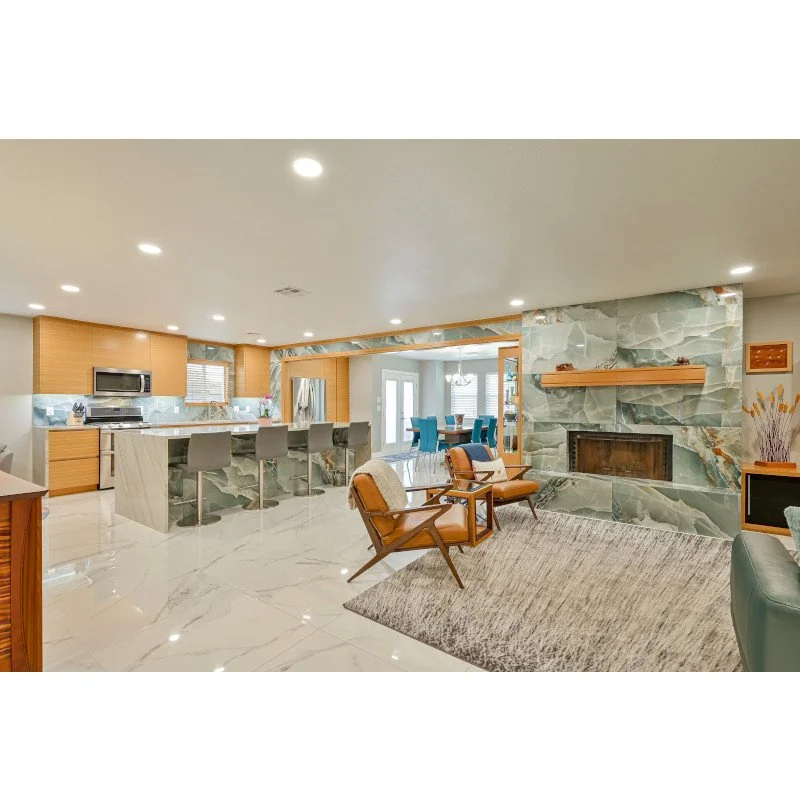 Open-concept living room and kitchen with marble flooring, a stone fireplace, tan chairs, and a kitchen with wooden cabinets and a marble island.
