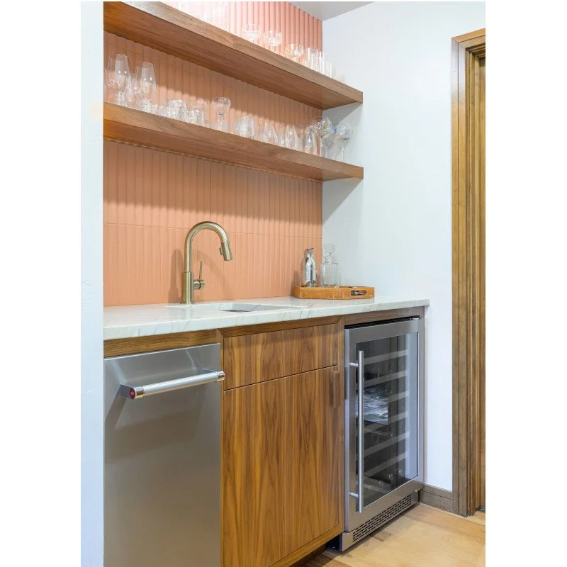 A small bar area with wooden shelves holding glassware, a countertop with a built-in sink, a small fridge, and a wine cooler, in a modern kitchen corner with a pink back wall.